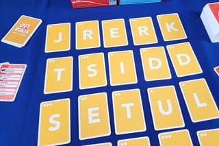 Wort-Ratespiel mit gelben Buchstabenkarten auf blauem Tisch bei der Queen-Games-Herbstneuheiten-Präsentation, im Hintergrund eine rote Queen-Games-Spielschachtel